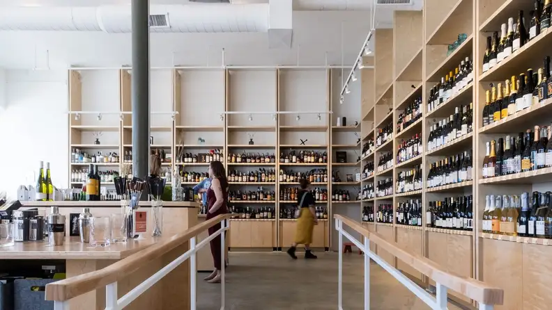 Four people shop for wine in a brightly lit natural wine store with light wood shelves filled with colorful bottles.