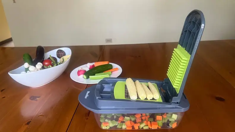A bowl and small saucer plate filled with assorted vegetables next to the Mueller vegetable chopper with 3 ears of baby corn on top of the slicer.