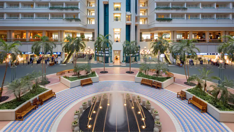 Spacious hotel atrium with indoor palm trees, a central fountain, and open seating areas surrounded by multiple floors of balconies and escalators.