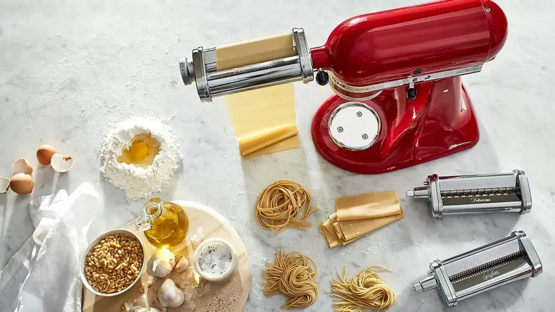 KitchenAid pasta attachment rolling pasta dough on a marble surface, surrounded by pasta ingredients