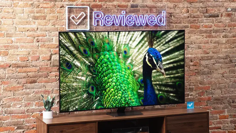 Samsung QN90D Mini-LED TV with vibrant peacock on screen atop a wooden TV stand below Reviewed neon sign in front of brick wall indoors.