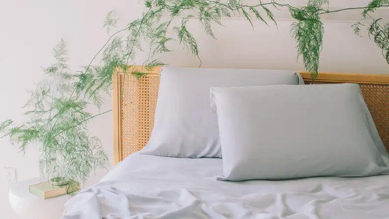 Wooden bed fitted with light grey sheets with plant on bedside table