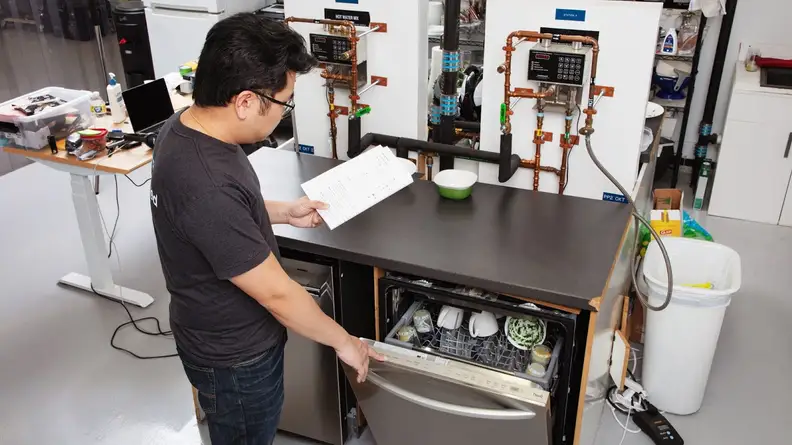 Our Senior Manager of Lab Operations, Jonathan Chan, stands before a dishwasher ready for testing in our labs, running through a checklist on a clipboard. Jonathan opens the dishwasher's door, showing an interior filled with dirty dishes.