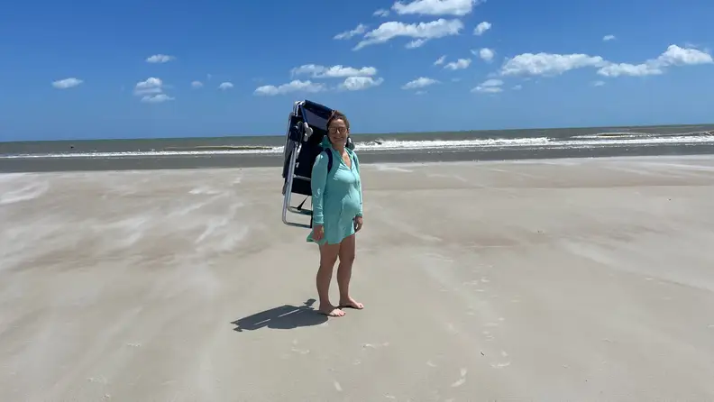 A woman standing on the sand wearing a backpack beach chair