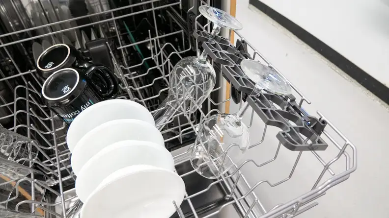 A close-up of the Maytag MDB4949SKZ top rack, focusing on its stemware holders. There are two wine glasses in the holders. The rest of the rack is full of clean bowls, mugs, and glasses.