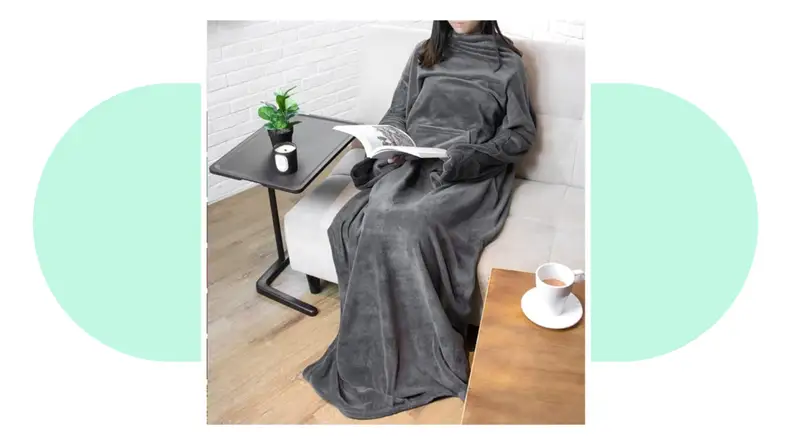 A person sitting down on a couch interacting with a laptop while wearing a fleece blanket over themselves.
