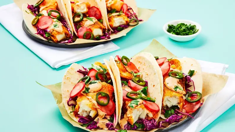 Three fish tacos arranged on a square white plate, accompanied by a second plate of tacos in the background.