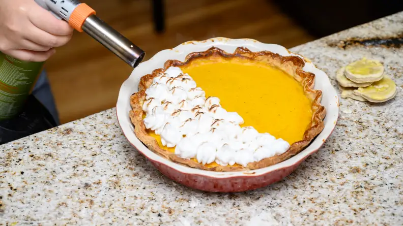 A hand using a butane torch to scorch meringue atop a pumpkin pie.