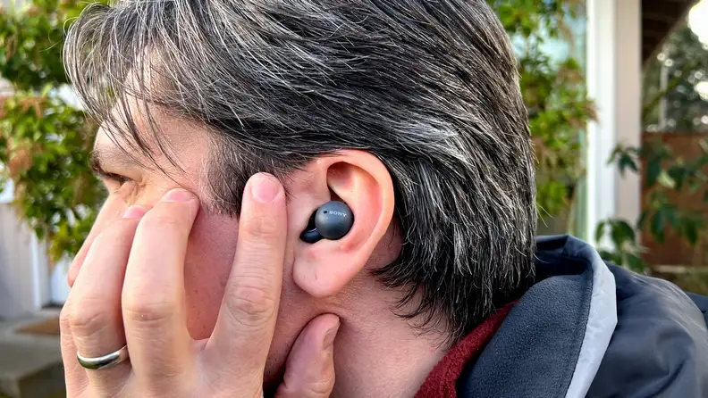 A man wearing a gray coat with salt and pepper hair taps his cheek in front of a grey, double looped earbud.