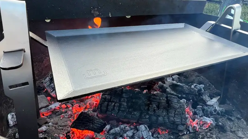 Made In Carbon Steel Griddle on top of open fire and smoky logs inside of grill.