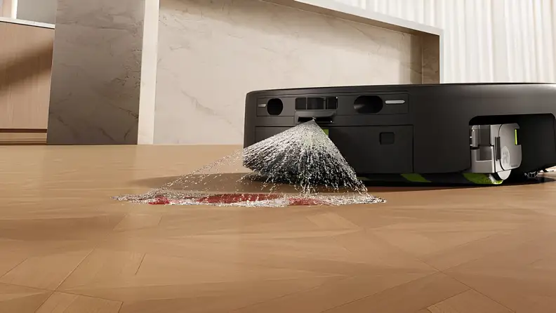 A close-up of an iRobot Roomba robot vacuum emitting a powerful high-pressure water spray onto a red stain on hardwood flooring, demonstrating its pre-treatment mopping technology.