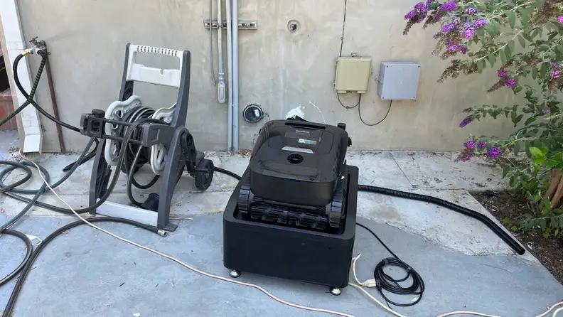Black Beatbot Aquasense X Robot Pool Cleaner next to house with hose and outlets next to it, beside a bush with green leaves and purple flowers.