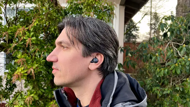A man wearing a gray coat with salt and pepper hair listens and smiles while wearing a double looped earbud in front of a bush.