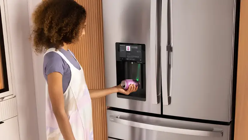 A person holds a beverage can under a smart refrigerator’s built-in dispenser in a modern kitchen.