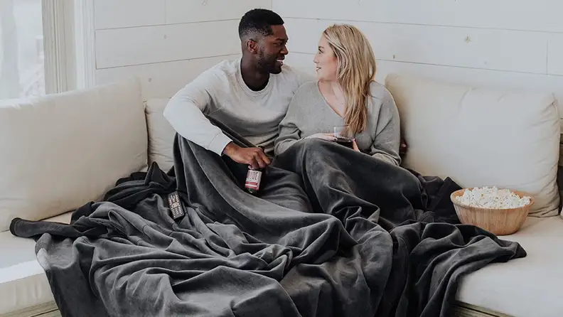 A couple lounges underneath a blanket inside.