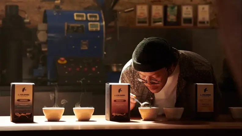 Person cupping coffee samples beside bags and a roaster