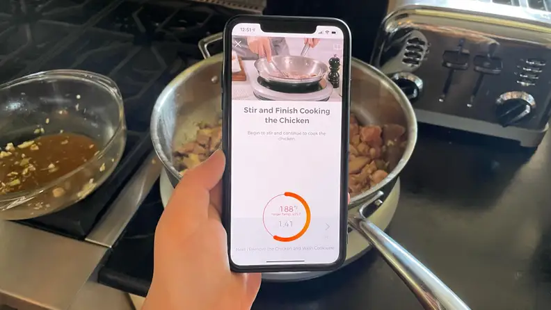 A person is holding a phone, which displays the guided cooking tutorial inside the Hestan Cue cooking app.
