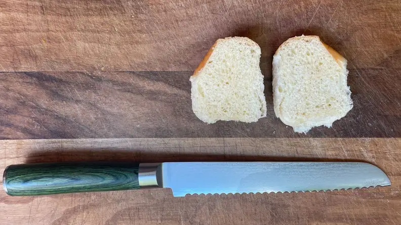 Jagged serrated knife from Hexclad's Damascus collection sitting below small roll of bread sliced in half on top of wooden cutting board.