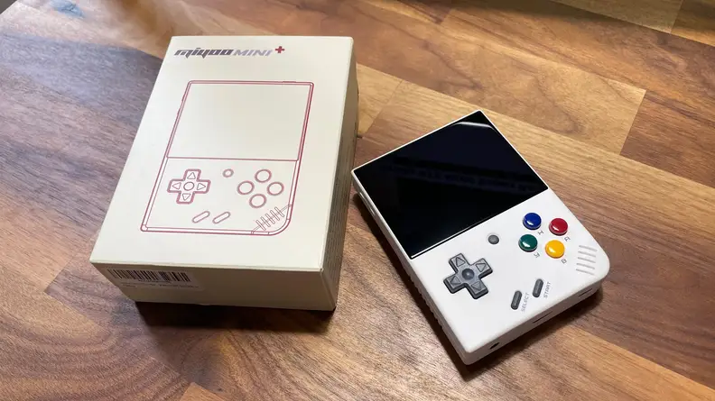 The Miyoo Mini Plus handheld console laid on a tabletop next to its beige, retro packaging.