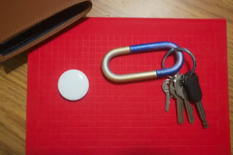 Apple's AirTag, on top of a red notebook, with a set of keys and a wallet nearby.