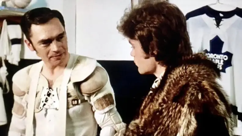 A man in hockey pads talking to another man in a fur coat.