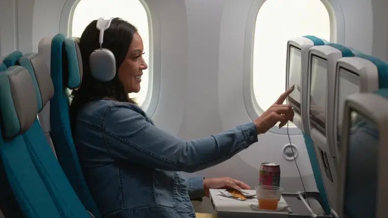 A woman in headphones taps the seatback screen on an Alaska Airlines flight, a drink and snack on her tray.