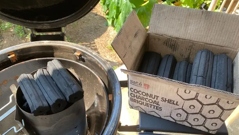 A box filled with charcoal briquettes sits next to a charcoal grill.