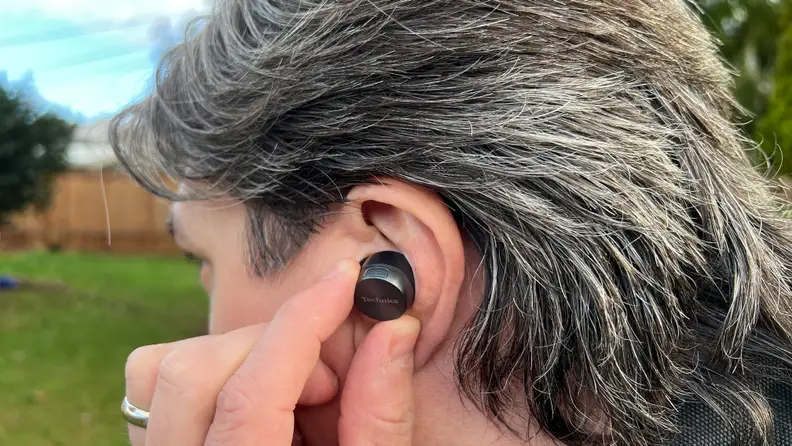 The gunsmoke colored wireless earbud sits in the ear of a man with brown and silver hair, with green grass and blue sky beyond.