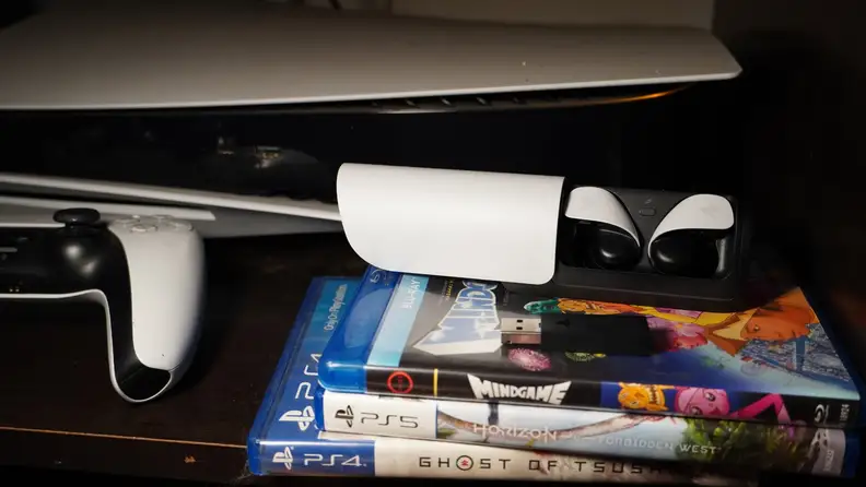 The earbuds case sitting atop a few PlayStation games with the DualShock 5 next to them.
