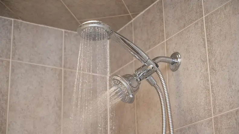 Handheld and permanent showerheads streams water in a beige-tiled shower stall.