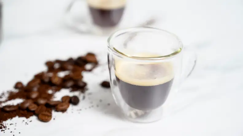 A clear espresso cup filled with freshly brewed espresso, accompanied by whole coffee beans scattered beside it.