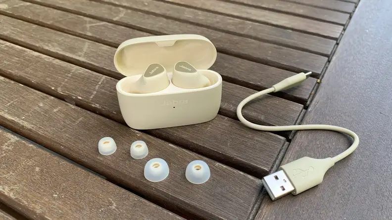 The gold beige Jabra Elite 5 in their open case with included USB-C to USB-A charge cable and extra ear tips on a wooden table.