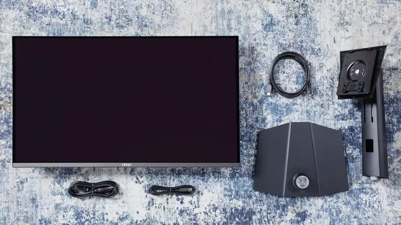 The The MSI MPG 321URX monitor and it's accessories laid out on a blue and white rug.