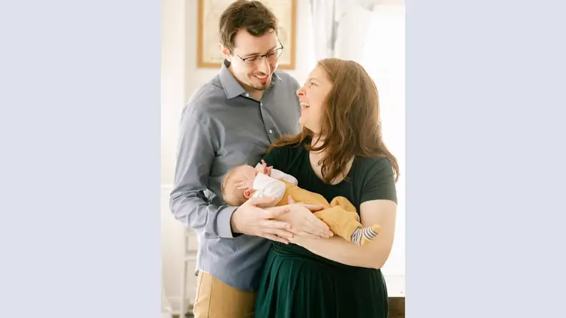 Two people smiling at each other while holding a newborn infant.