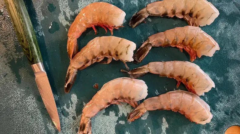 Small utility knife with green knife sits next to 7 pieces of raw pieces of raw shrimp on countertop surface.
