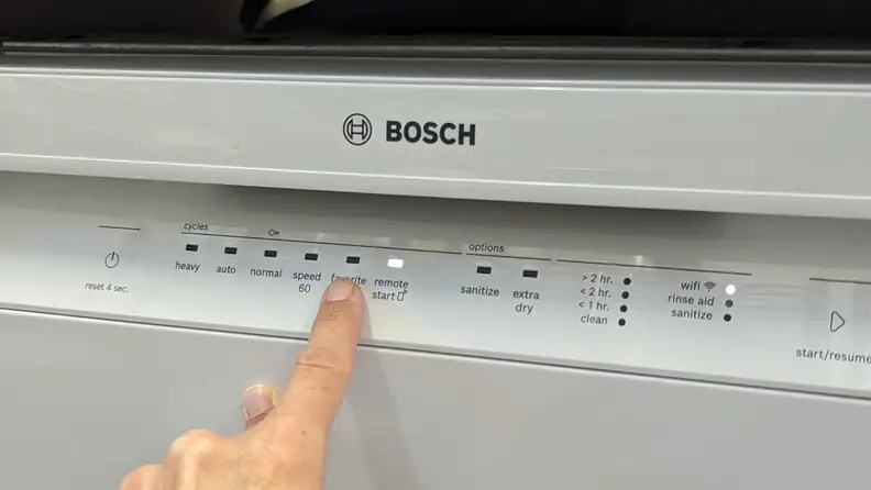 A finger points to the "Favorite" cycle button on the front-facing control panel of the Bosch 100 Series SHE3AEM2N dishwasher.