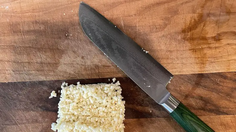 Hexclad's Damascus chef's knife with green handle next to square pile of rice on top of wooden cutting board.