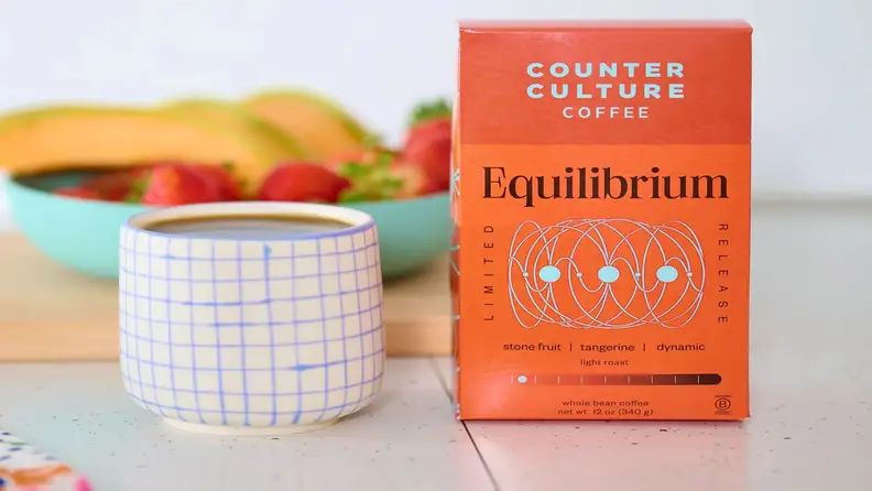 Coffee box beside a mug with fruit bowl in background