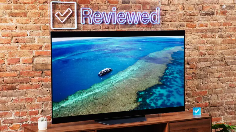 The LG B2 on a wooden table in front of a brick wall with a neon Reviewed sign displaying a beach.