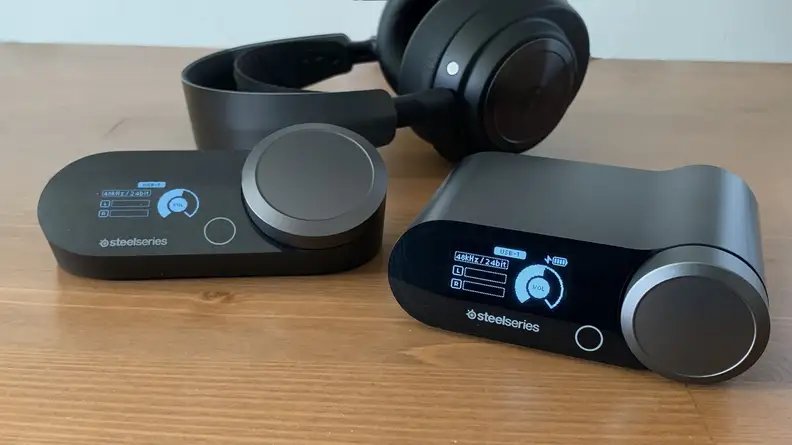 An Arctis Nova Pro wireless and wired base station sit on a wooden table in front of a headset.