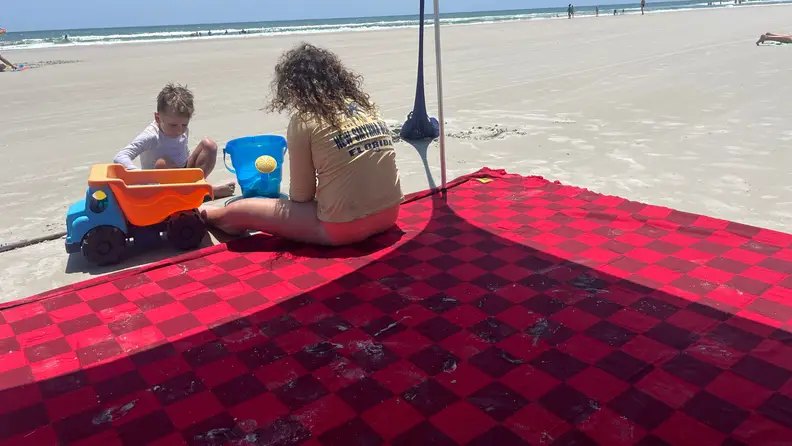 Two small children playing on the edge of red and burgundy Big Beachy blanket on sand at beach.