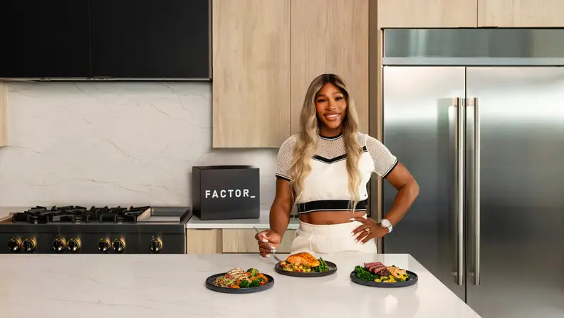 Serena Williams stands in a kitchen with a Factor box on the counter, plating three Factor meals in front of her.