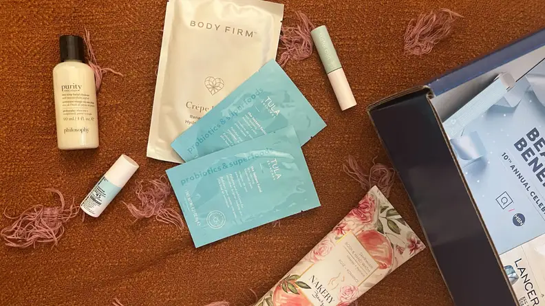 An image of several different makeup and skincare products on an orange bedspread, including a purity skin cleanser, three cleansing masks, a Nakery Beauty rose-scented body and hand wash, and a Tarte Sea mascara tube.