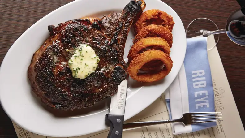 Ribeye with steak butter on top, onion rings, and branded knife, all on a white oblong plate