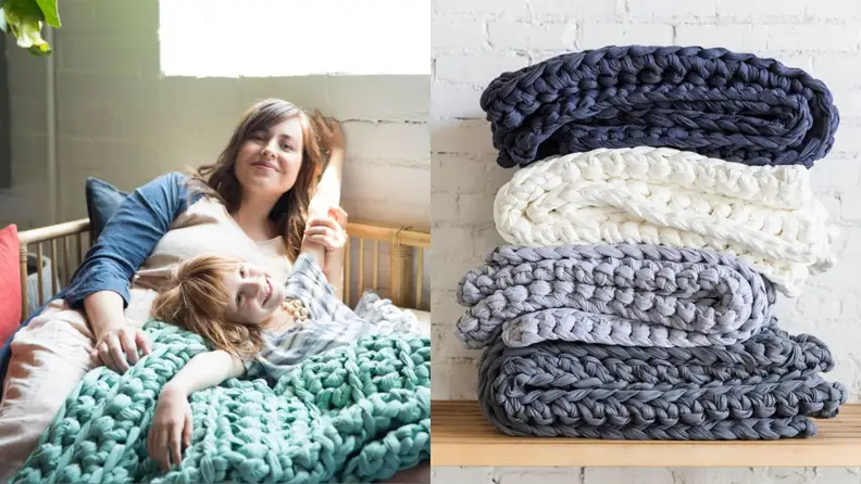 A mom laying with her daughter, wrapped up in a cozy, crochet blanket. A stack of colorful blankets in shades of blue.