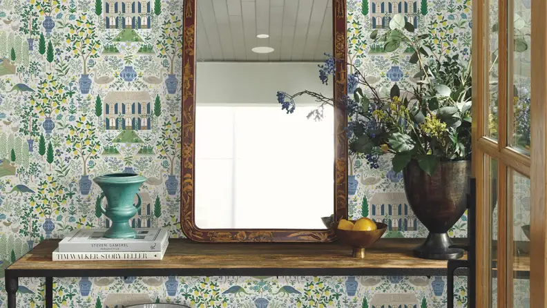 White, blue, yellow, and green wallpaper featuring a small vintage house next to fig and lemon trees. A wooden table with a mirror sits in front of the wallpaper