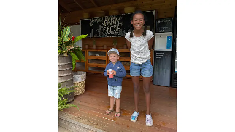 Two children smiling outside a farm stand with fresh produce and drinks.