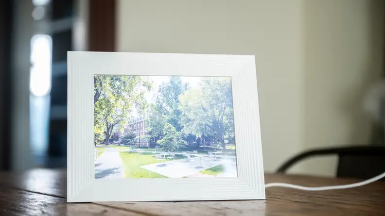 A white Aura digital picture frame displays an image of a brick building surrounded by trees.
