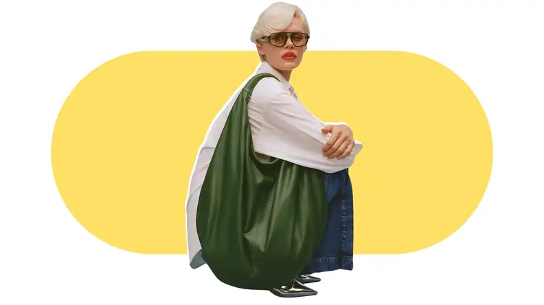 A model holding a very large green leather tote bag that covers her crouching body.