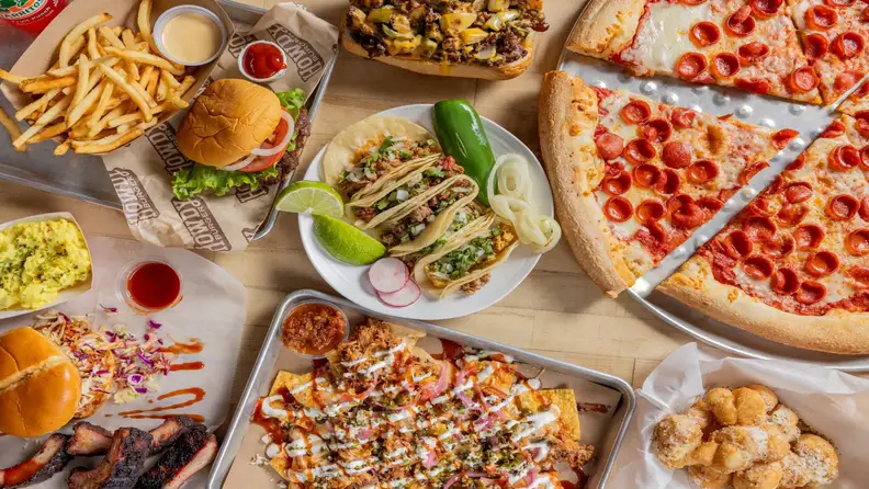 Overhead spread of pizza, tacos, burger, cheesesteak, and nachos